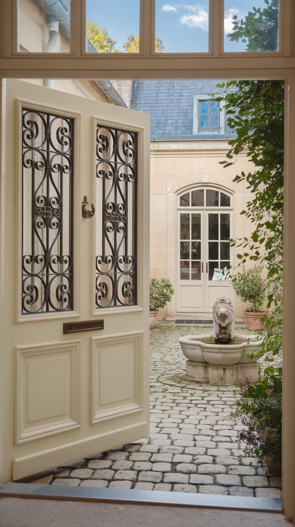 A cream front door with wrought iron details leading to a cobblestone courtyard with a fountain and potted plants.