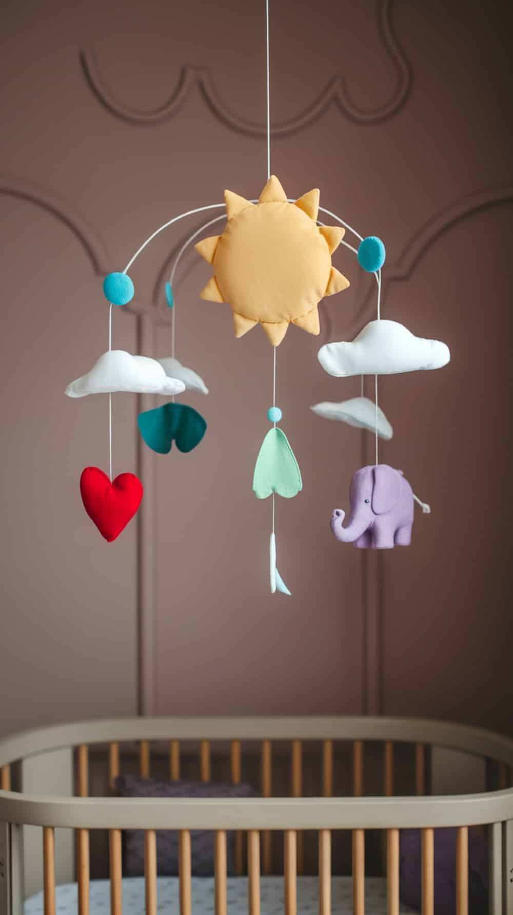 A colorful nursery mobile featuring a sun, clouds, a heart, and an elephant, hanging above a crib in a taupe nursery.
